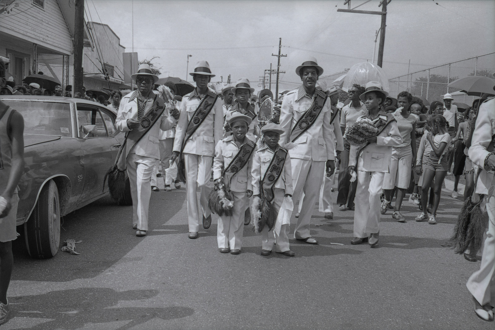 Norman Dixon, Sr. parading with the YMO, Jr., 1977. A group of men and children dressed in matching white suits, hats, and sashes march down a street during a second-line parade. They carry feathered fans and bouquets, surrounded by a crowd of onlookers holding umbrellas.