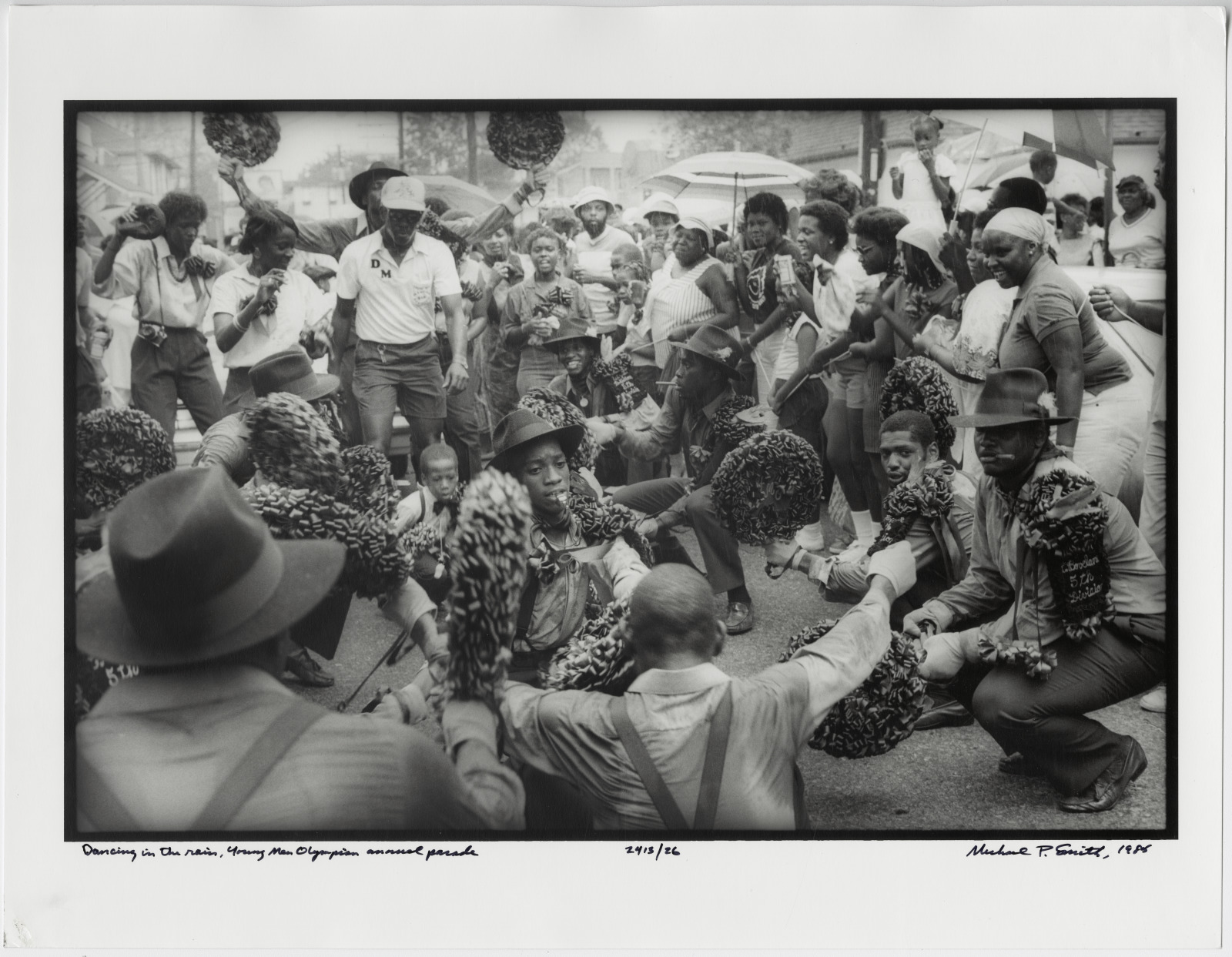 In a black and white 1985 photo, a group of dancers in matching outfits crouch in a circle, holding decorated fans, as a lively crowd watches during the Young Men Olympian annual parade.