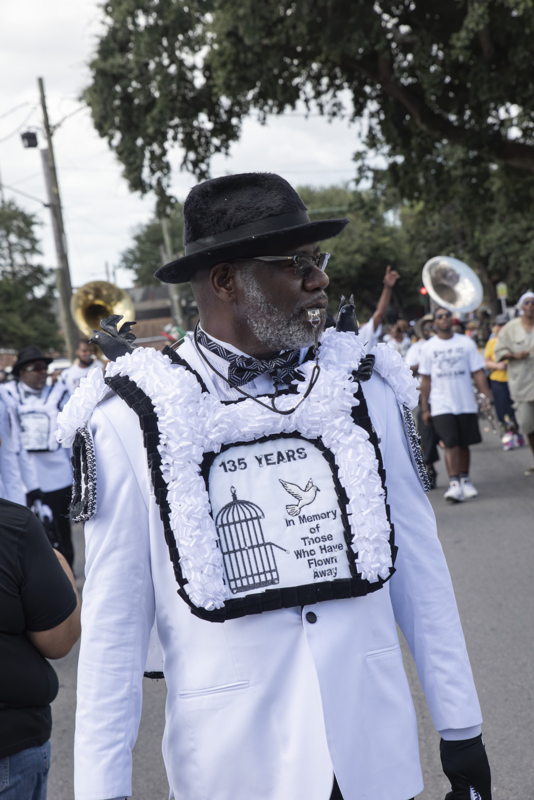 Norman Dixon Jr., President of the YMO Jr., walks down the street at a parade with a whistle between his lips. He's wearing a crisp white suit, black hat, and a decorative chest piece commemorating the group's 135 year anniversary.