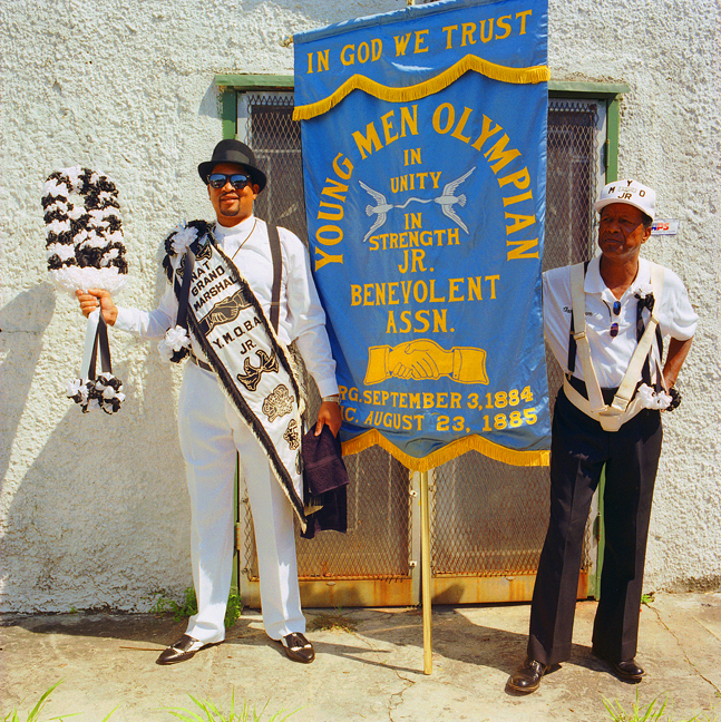 Grand Marshal Joseph Spots stands next to banner holder Charlie Brown, in front of a white stucco building. Spots wears a white suite with a decorative sash.