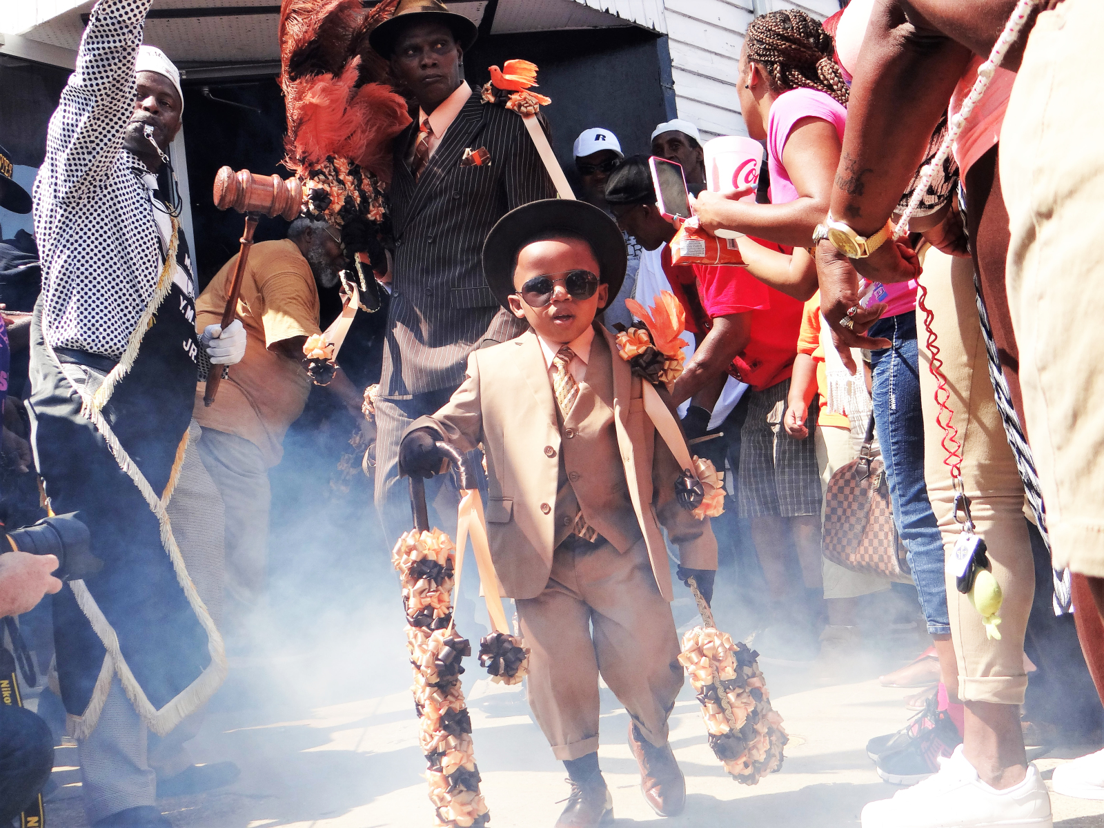 A child from The New Look, YMO, Jr.'s youth division, comes out a door at the beginning of a second-line parade in 2016. He is wearing a suit and hat with dark sunglasses.
