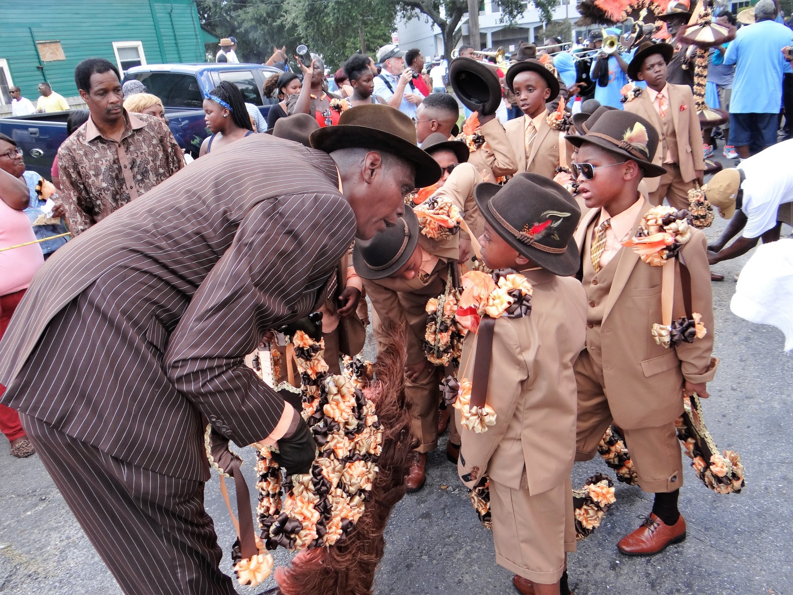 Jack Humphrey leans over to talk with a group of children at a second-line parade, who are part of The New Look, YMO, Jr. youth division. They are wearing green suits and hats with sunglasses.