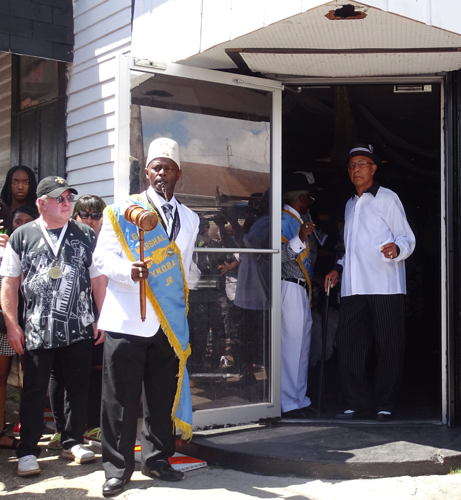 Quint Davis, Jerome “DJ Jubilee”, and Richard Martin stand near the open doorway of YMO Jr.'s building.
