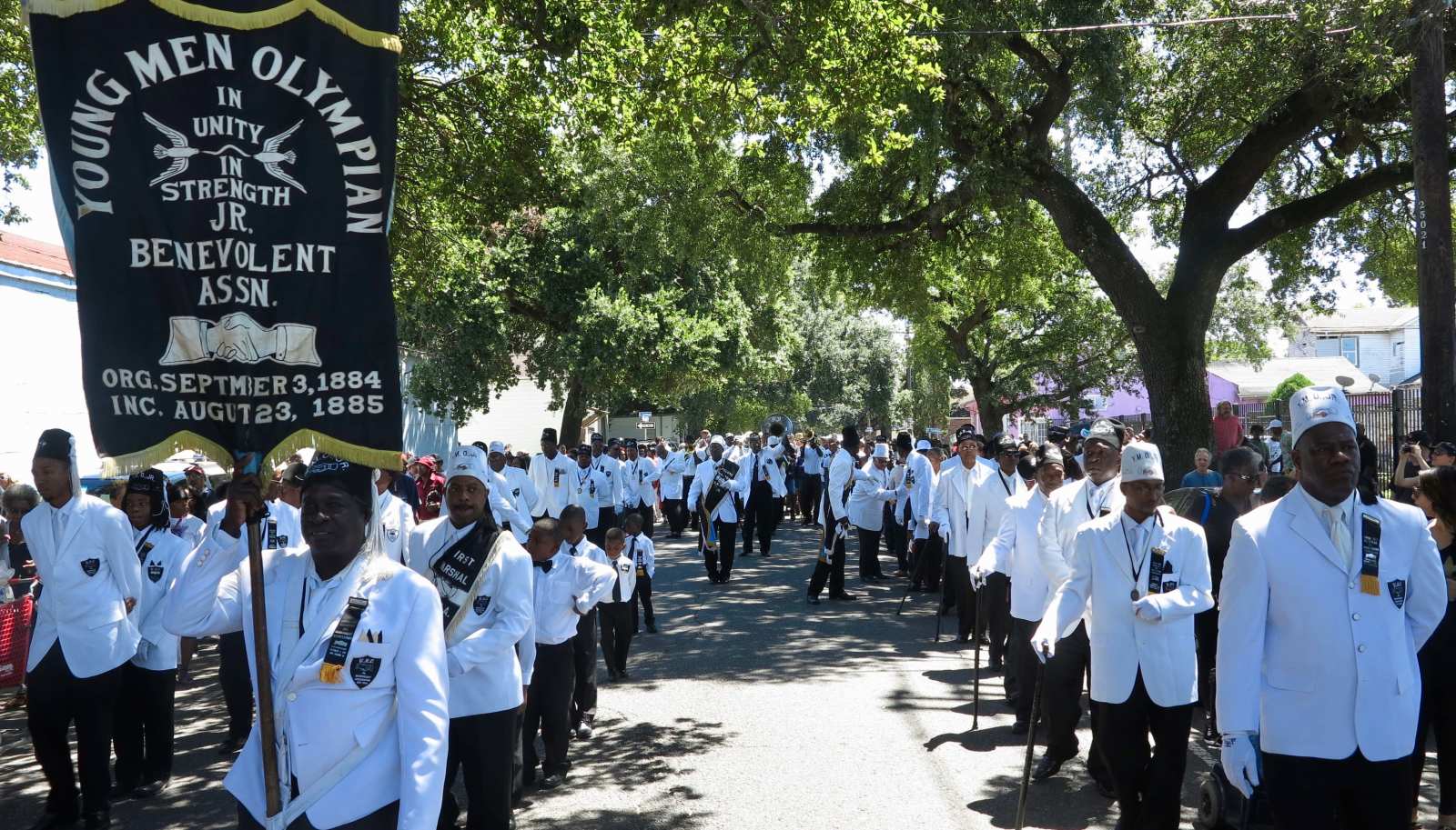 A procession of men in matching white suits process down a street during YMO Jr.'s anniversary parade on a sunny day. One of them holds a banner that reads "YOUNG MEN OLYMPIAN Benevolent Assn."