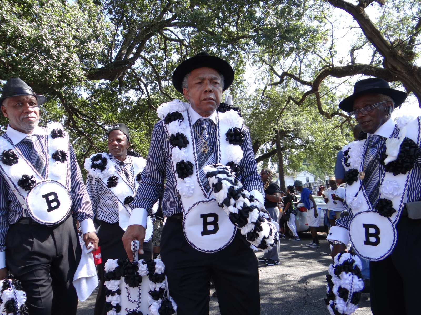 Three men, who are part of The Body at a YMO Jr. parade gather on the street. They area wearing matching suits with white sashes that that hold round emblems marked with the letter "B".