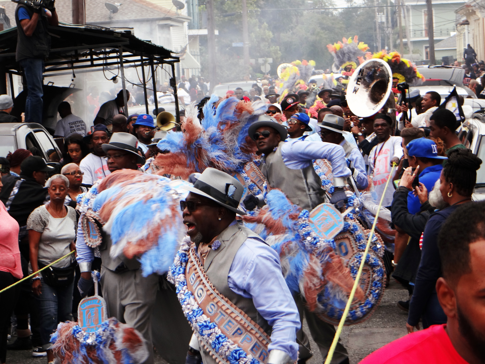 The Big Steppers dance during a YMO Jr. parade. They are wearing matching blue and green suits and are followed by a brass band and a crowd, flanked by food vendors.