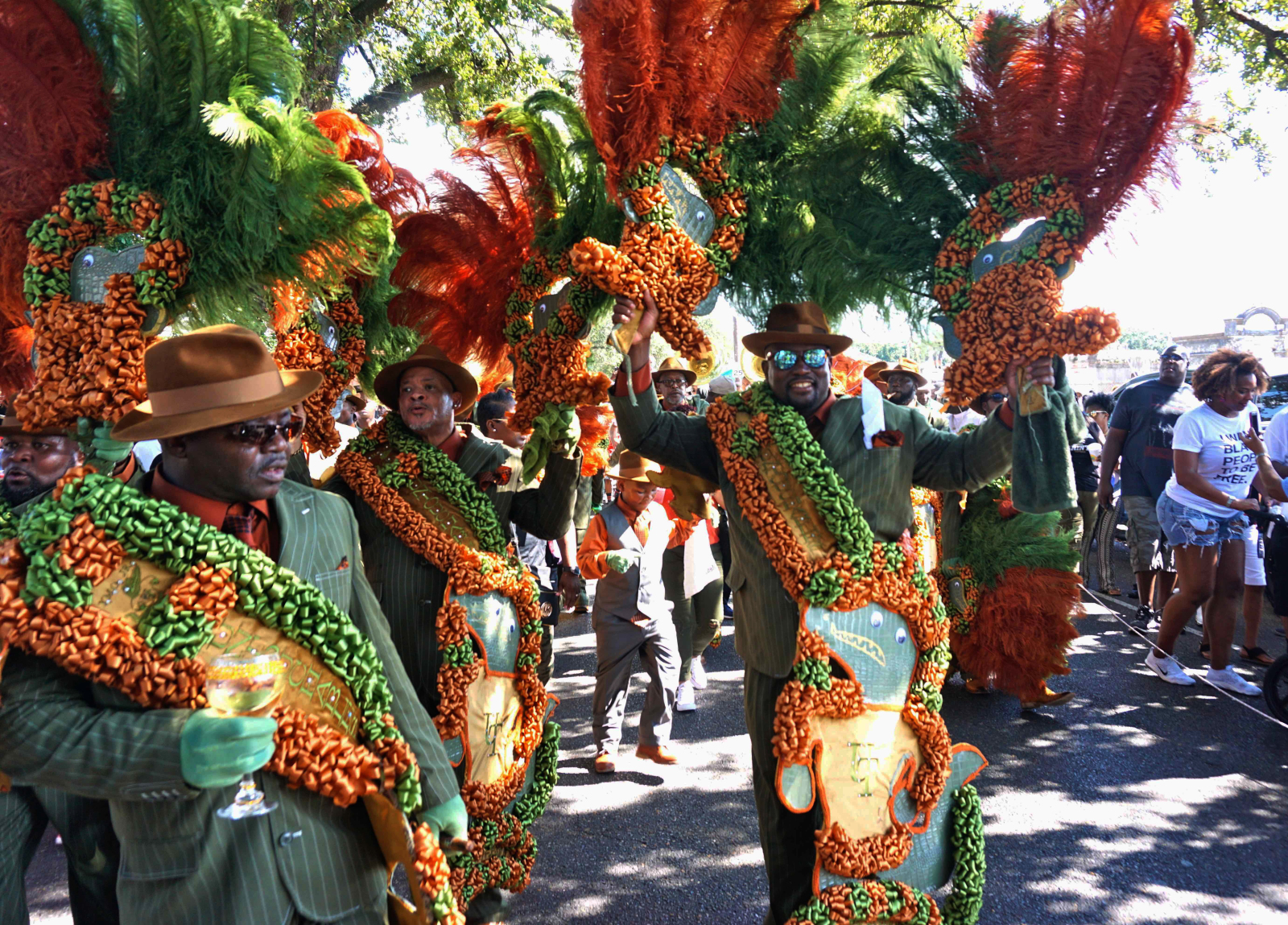 Several men who are part of The Untouchables walk down the street with greena nd yellow feathered streamers and green suits at a YMO Jr. parade.