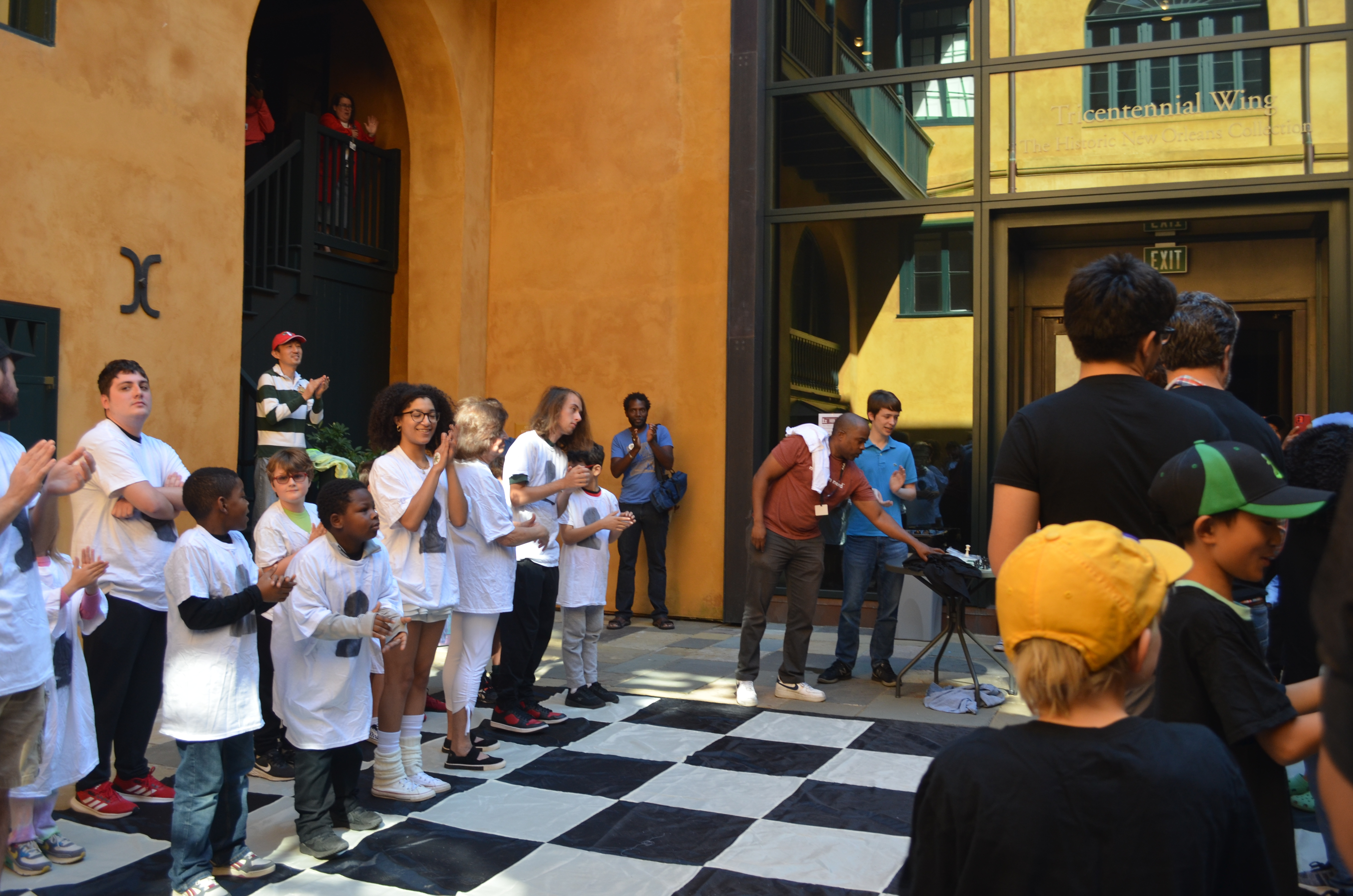 A diverse group of people, including children and adults, stands on a large checkered floor in an outdoor courtyard. Theyre watching a person speaking into a microphone. The building is yellow with green accents. Others are clapping and observing.