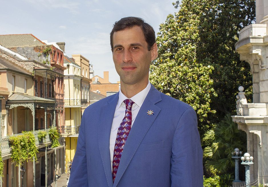 A man in a blue suit and patterned tie stands outside. Behind him is a row of colorful historic buildings and a large tree. The sky is clear, and the setting appears to be a picturesque urban area.