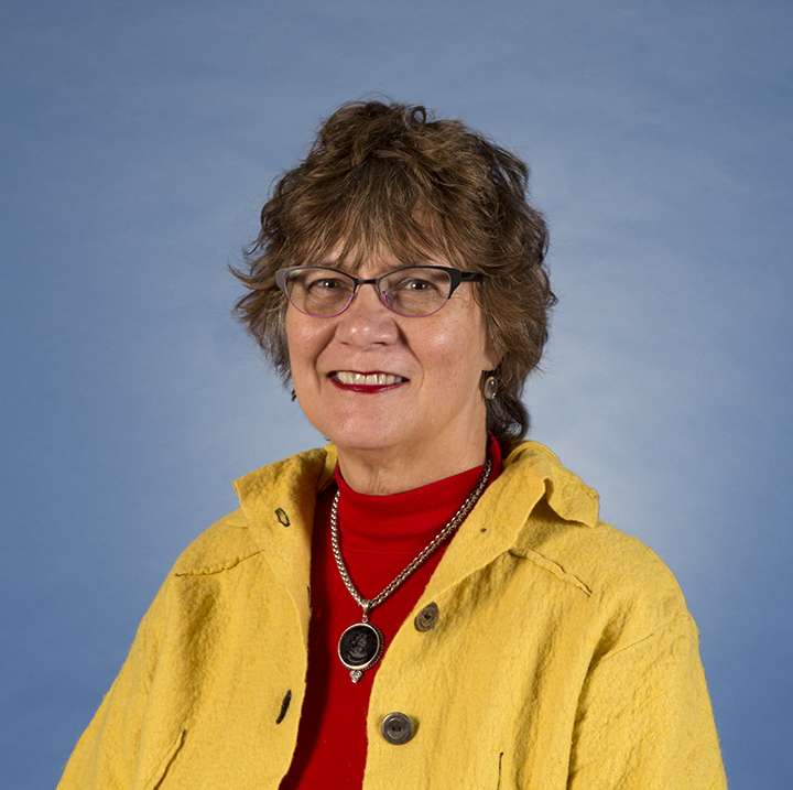 A person with short, curly hair is smiling while wearing glasses, a yellow jacket over a red shirt, and a necklace. The background is a solid blue color.