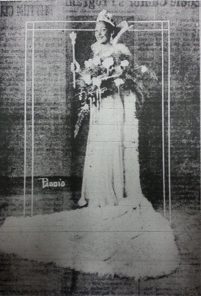 A black and white vintage photo of a person dressed in an elegant gown, holding a bouquet and a scepter. They are wearing a crown and a fur-trimmed cape, standing on a textured surface with a backdrop of printed text.