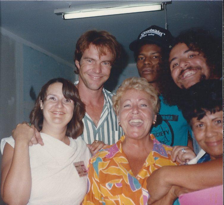 A group of six smiling people pose closely together indoors. They appear to be in a casual setting, with overhead lighting. The individuals show a mix of casual and colorful clothing, and their expressions suggest they are enjoying each others company.