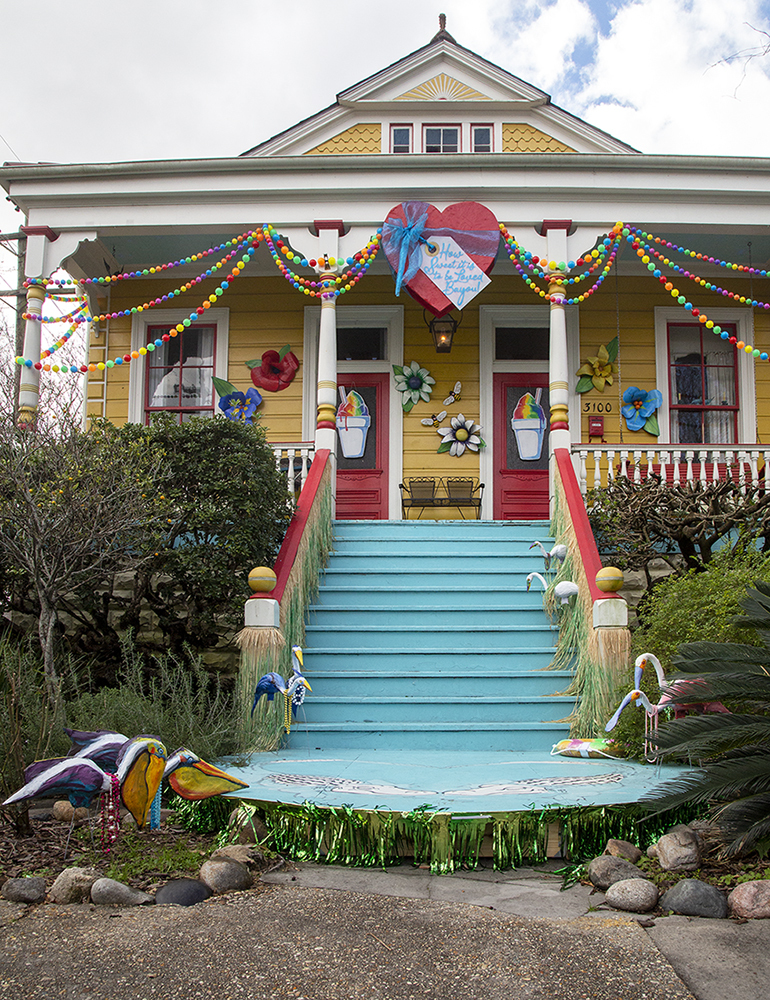 A colorful yellow house decorated with beads, flowers, and garlands. The front stairs are painted blue, flanked by green fringe and decorative pelicans. The porch features banners and a heart-shaped sign, celebrating a festive, cheerful atmosphere.
