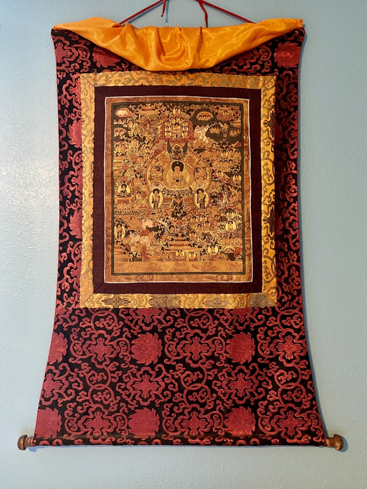 A colorful tapestry hangs on a light blue wall. It features an intricate design with a central golden figure surrounded by detailed patterns and smaller figures. The tapestry has rich red, black, and gold tones with an ornate border.