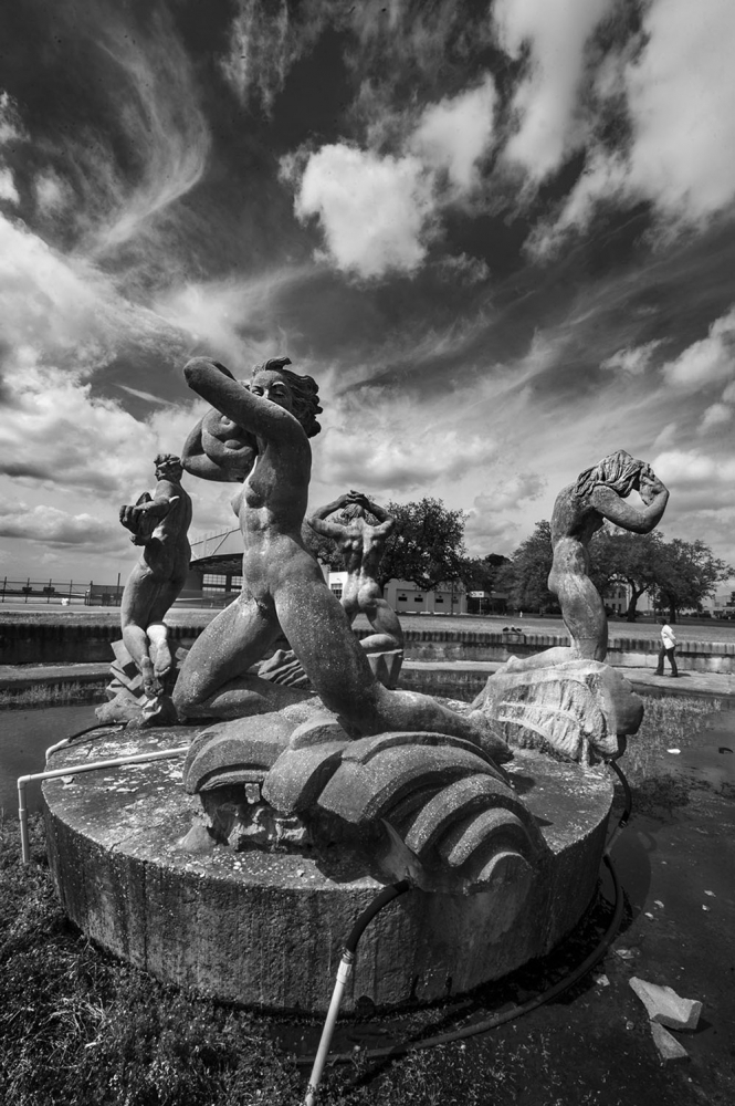 A black and white photo of a dynamic stone sculpture featuring three mythical figures in dramatic poses atop waves, set against a sky filled with swirling clouds. A person stands in the background near the waters edge.