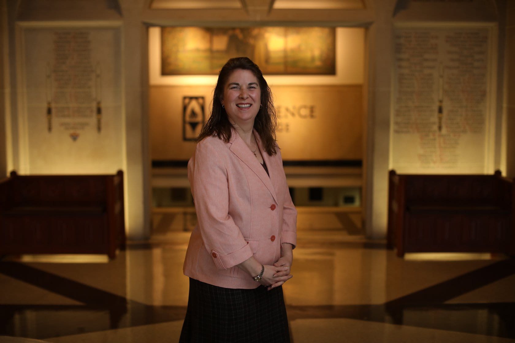 A woman in a pink blazer stands smiling in a grand hallway with polished floors. The background features an illuminated wall with inscriptions and wooden benches on either side.