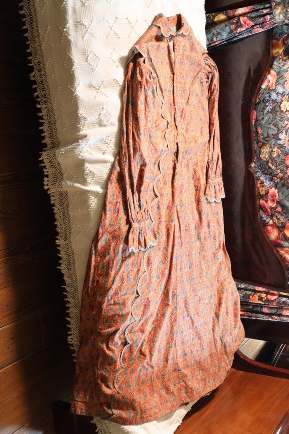 A vintage orange long-sleeve dress with a paisley pattern hangs against a textured curtain. The dress features a high collar, decorative scalloped trim, and ruffled cuffs. Floral and wooden backgrounds complement the antique aesthetic.