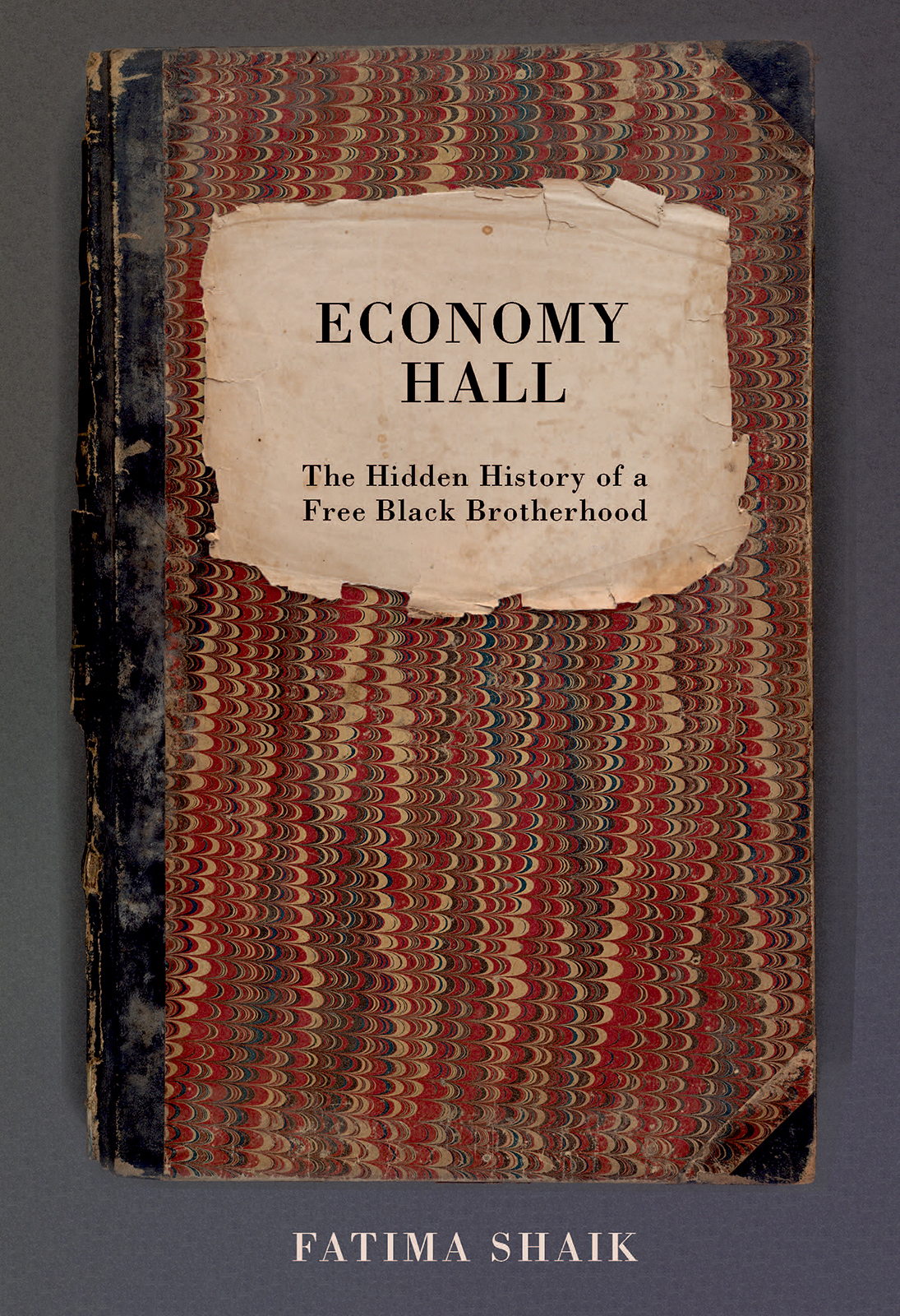 The image shows a book titled Economy Hall: The Hidden History of a Free Black Brotherhood by Fatima Shaik. The cover features a vintage, worn notebook with a marbled design and a paper label.