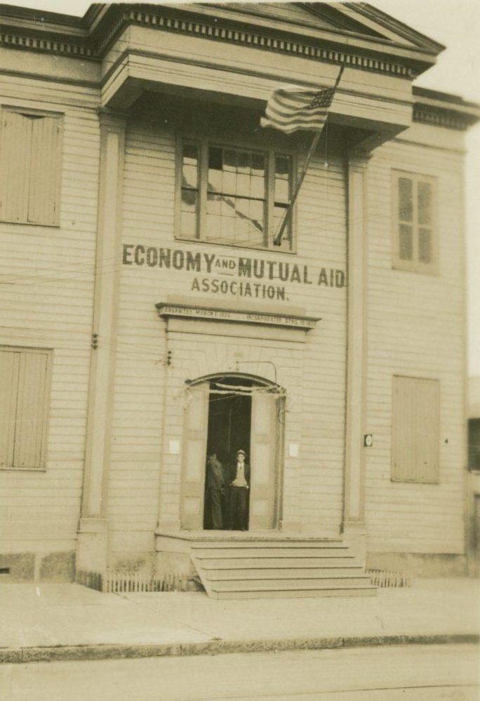 A vintage photo of a two-story wooden building with Economy and Mutual Aid Association written above the entrance. Two people stand in the doorway, and an American flag is flying above. The windows are shuttered.
