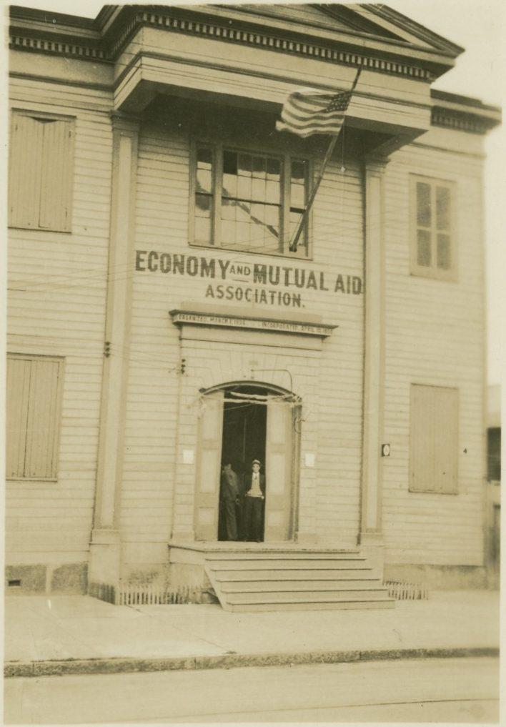A sepia-toned photo of a two-story wooden building with an American flag above the entrance. The sign reads Economy and Mutual Aid Association. Two people stand in the open doorway on a quiet street.