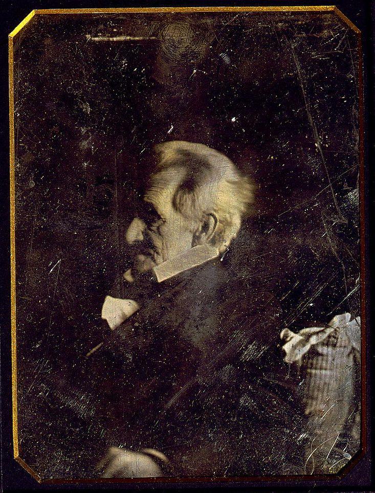 A vintage photograph of an elderly man in profile, sitting and appearing contemplative. The image has a dark, worn appearance with visible scratches and a golden border, creating an antique feel.
