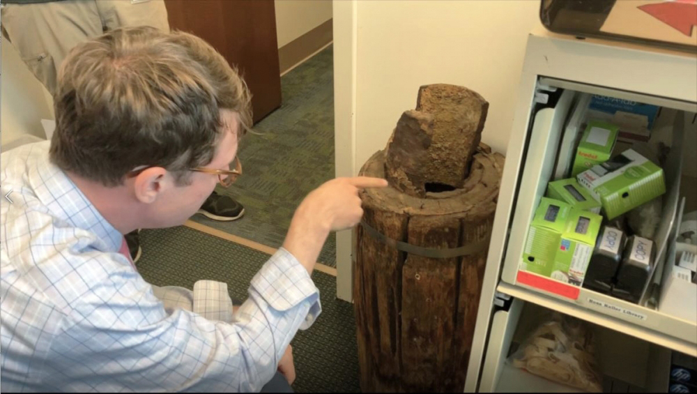 A man wearing glasses and a plaid shirt points at a large, rusted metal canister sitting on the floor. The canister has a damaged, rusty top. To the right, a shelf holds various small boxes and a jar.