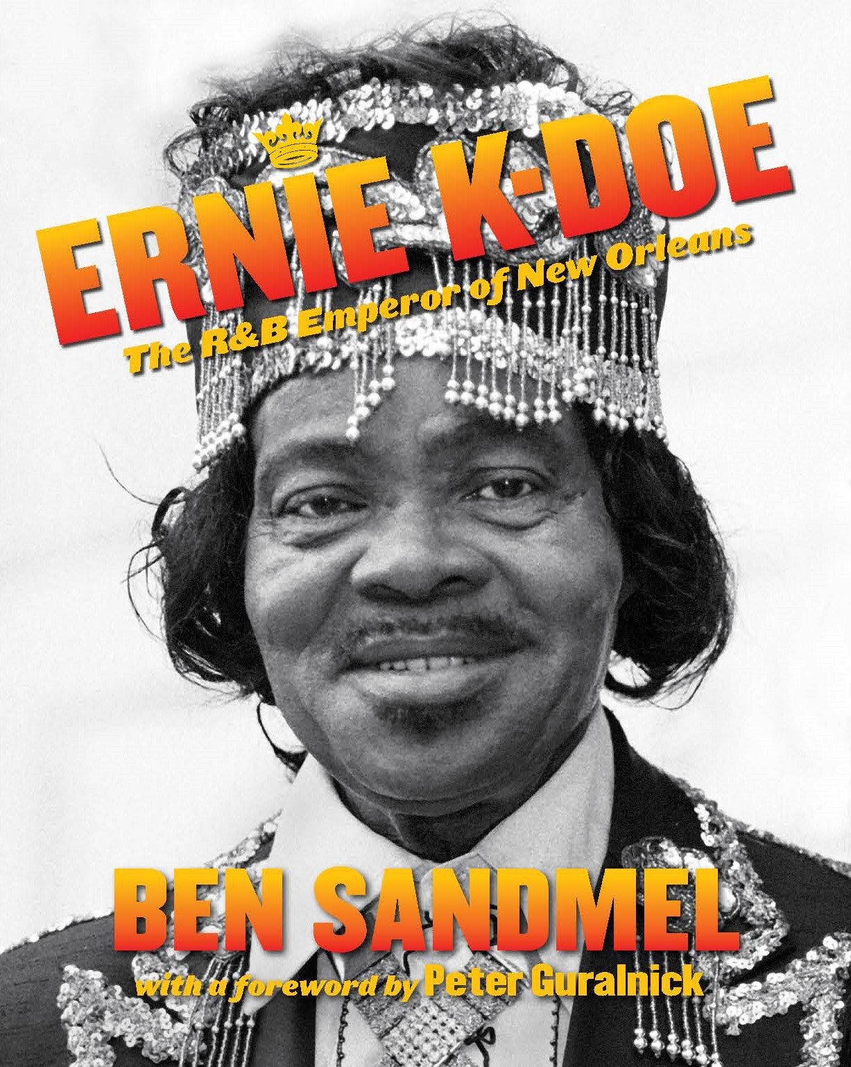 Black and white photo of a person wearing a crown and ornate clothing, smiling. The text reads Ernie K-Doe: The R&B Emperor of New Orleans by Ben Sandmel, with a foreword by Peter Guralnick.