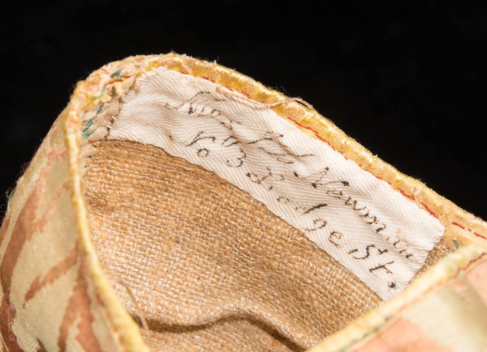 Close-up of a woven fabric with a handwritten label stitched inside. The label appears to have text that is partially visible, with a date and possible location or event details. The fabric is textured with a beige and orange pattern.