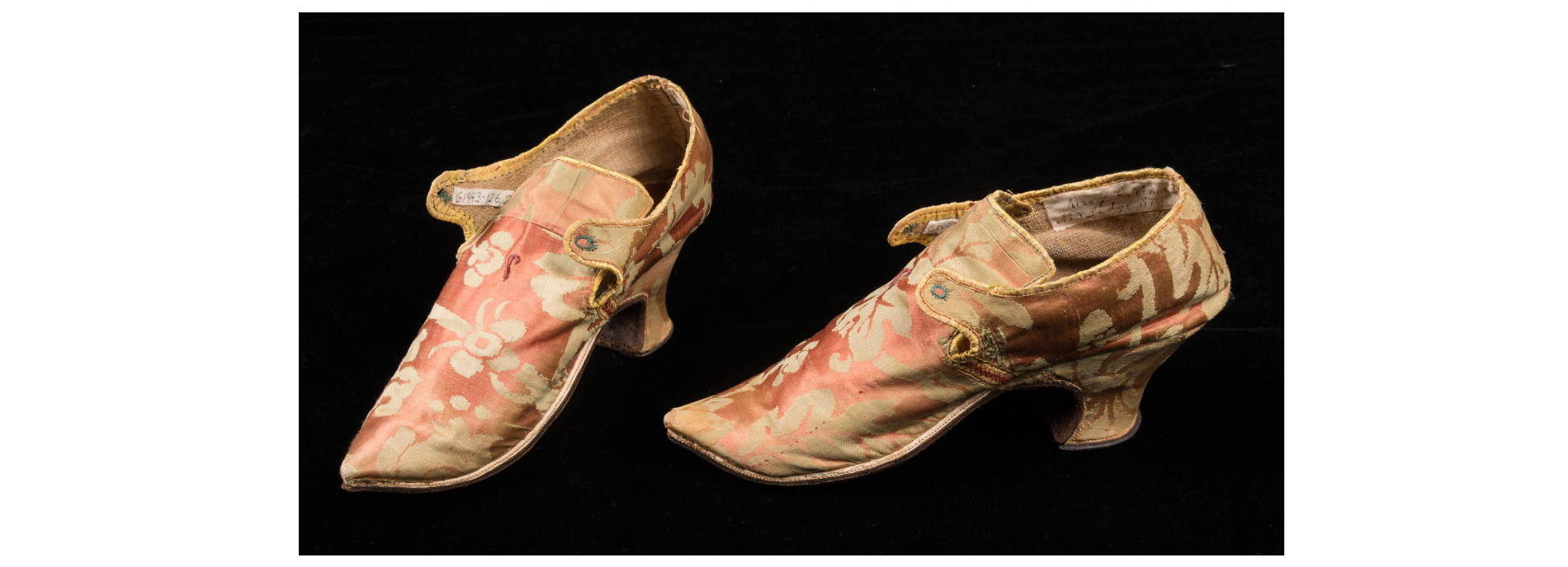 A pair of vintage floral-patterned shoes with pointed toes and small heels. The fabric is a soft pink with cream and yellow designs. The shoes have a strap with a button on the side and a slightly worn appearance.
