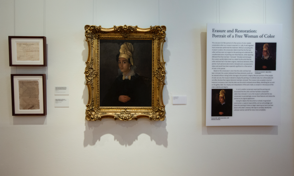 A museum exhibit featuring a framed portrait of a woman wearing a head wrap, flanked by two documents on the left and an information panel on the right titled Erasure and Restoration: Portrait of a Free Woman of Color.