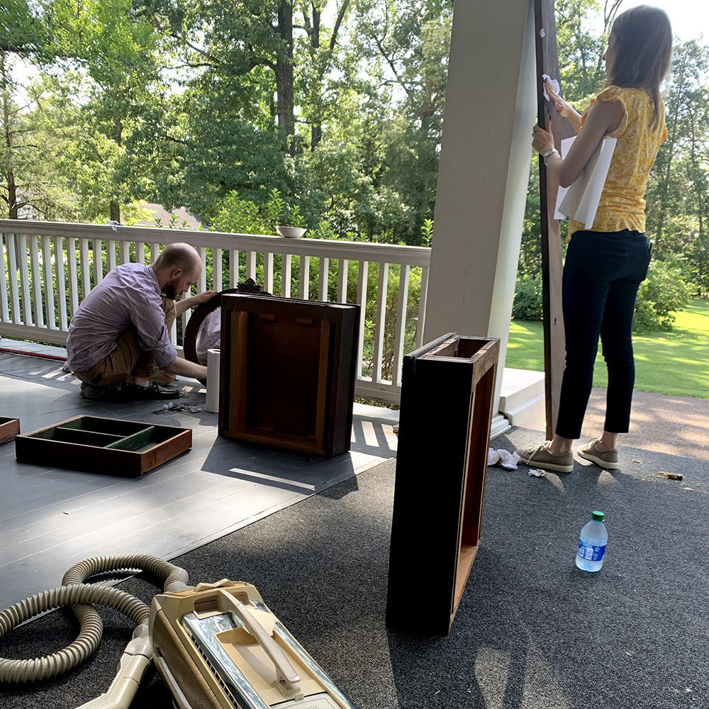 A man kneels on a porch assembling furniture pieces, while a woman stands nearby checking a part. Various wooden components and tools are scattered on the ground. The porch overlooks a lush, green yard.
