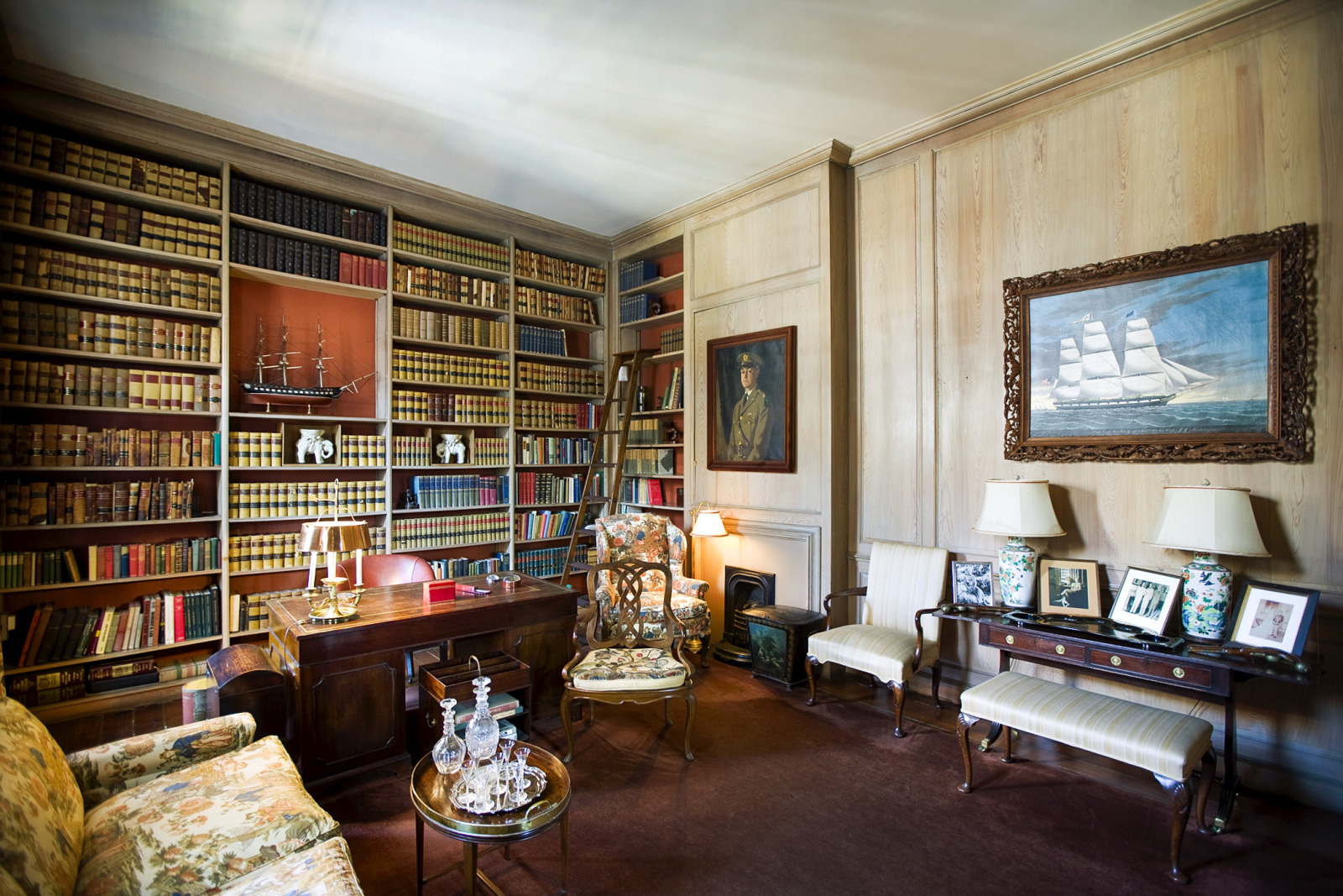 A cozy, elegant library room with wooden bookshelves filled with books, a wooden desk with a green lamp, decorative ship models, and framed paintings on the walls. Two white chairs and a floral sofa flank a small glass-top table.