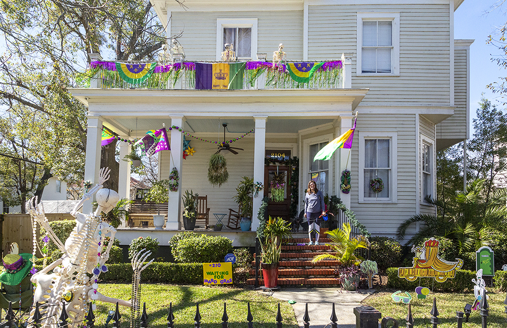 A two-story house decorated with colorful Mardi Gras banners, beads, and skeletons. A person stands on the porch steps surrounded by vibrant plants and festive decorations. A small dog sits nearby. Lush greenery borders the yard.