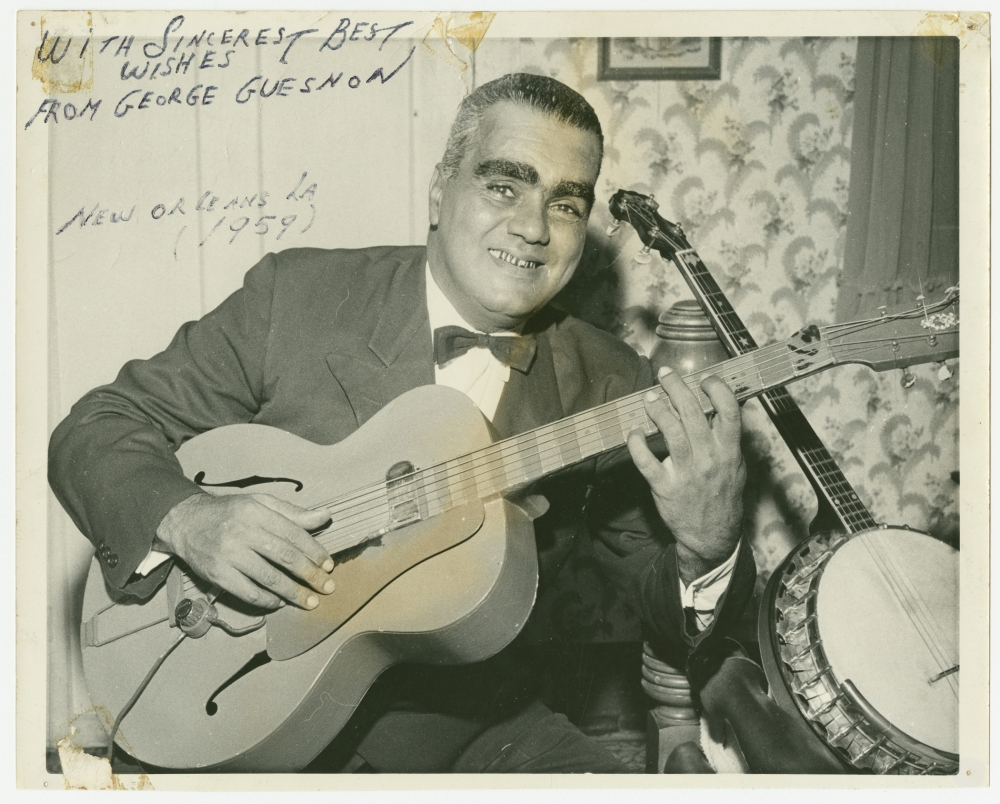 A man in a tuxedo smiles while holding a guitar. A banjo rests beside him. The handwritten caption in the corner reads With sincerest best wishes from George Guessnon, New Orleans, La (1959). The background features patterned wallpaper.