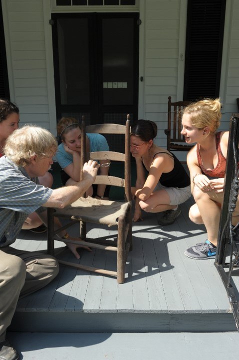 A group of five people gather around an old wooden chair on a porch. They appear to be examining or discussing the chair closely. The porch has gray wooden flooring, and there is a building with white siding in the background.