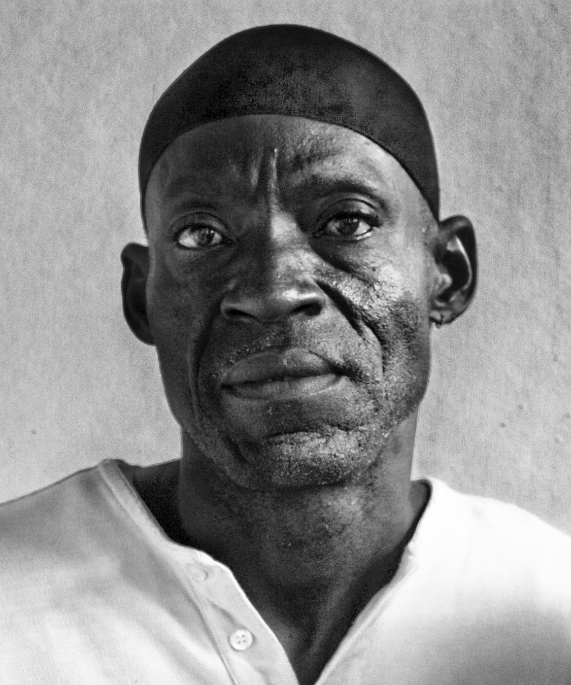 A black and white portrait of a man wearing a dark headwrap and a light-colored button-up shirt. He gazes directly into the camera with a neutral expression, against a plain background.