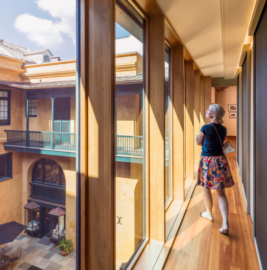 A person with gray hair stands in a glass-walled corridor, overlooking a sunlit courtyard. The interior features wooden panels and an elegant, historic architectural style.