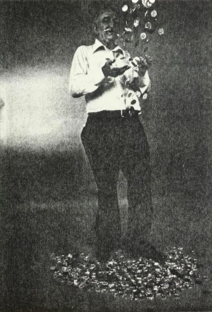 A person stands on a pile of bottle caps, looking down as more caps fall around them, holding some in their hands. The background is a blurred gray. The scene has a vintage, high-contrast appearance.