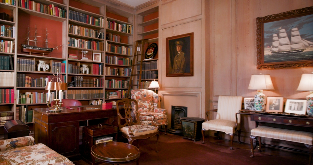 A cozy library with wooden bookshelves filled with books and decorative items, including model ships. Floral armchairs, a wooden desk, and lamps add warmth. Paintings of ships and a portrait adorn the walls, creating an inviting reading space.