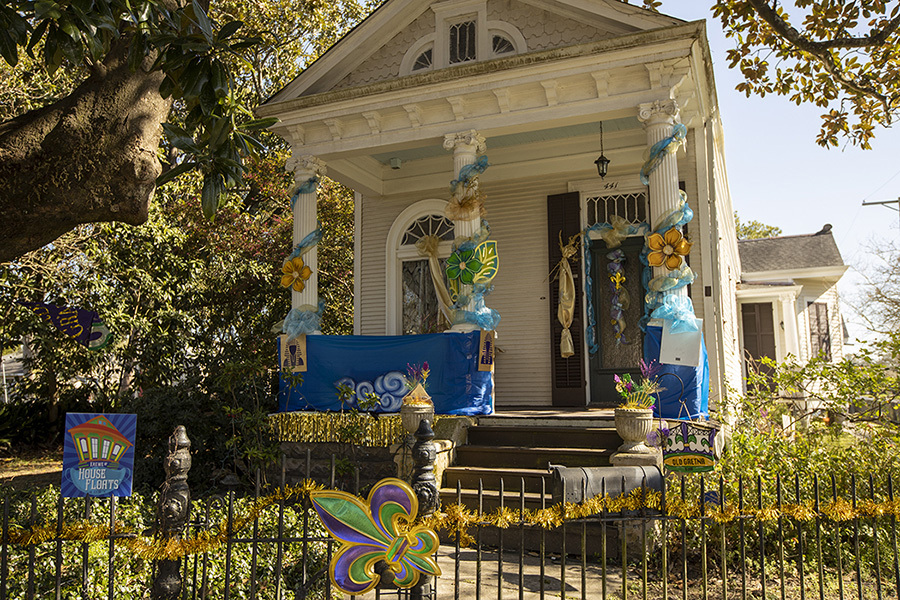 A festive house decorated with colorful Mardi Gras-themed banners, masks, and flowers. The house features a small porch with blue and gold streamers, and a fence adorned with a tinsel garland in the front yard.