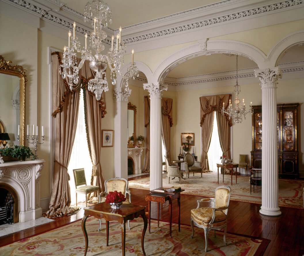 Elegant room with ornate chandeliers, tall columns, and large windows with draped curtains. Antique furniture, including chairs and tables, are arranged on a patterned rug over polished wood floors. A fireplace and grand mirrors add to the classic decor.