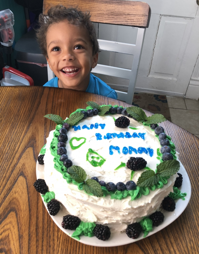 A smiling child sits at a table with a homemade cake in front. The cake is decorated with blueberries, blackberries, and mint leaves, with Happy Birthday Mommy written in blue icing.