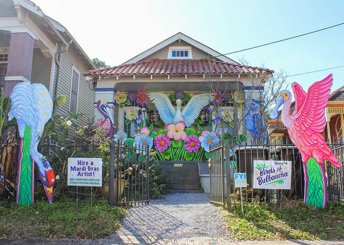 A house adorned with vibrant Mardi Gras decorations, featuring large, colorful birds and flowers at the entrance. Signs promote Mardi Gras artists and decorations. The scene is lively and festive, set in a residential neighborhood.