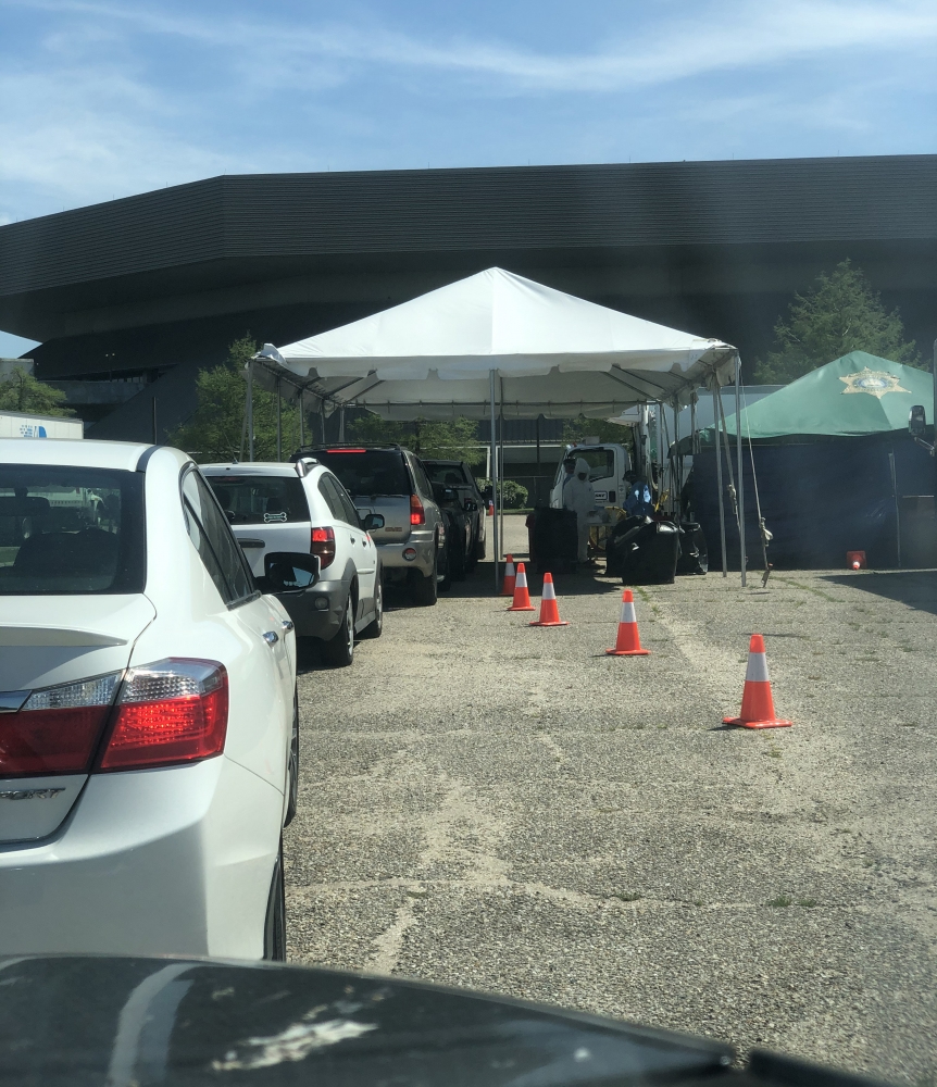 Cars are lined up at a drive-through testing site. White tents provide shade for workers in protective gear. Traffic cones guide the vehicles. A large building is visible in the background.