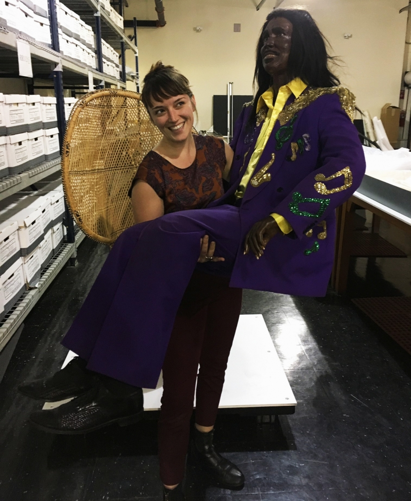 A woman is smiling while holding a life-sized mannequin dressed in a purple suit with gold embellishments. They are in a room with shelves of boxes in the background.