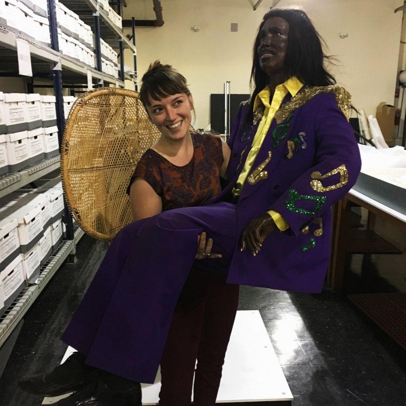 A woman is smiling while holding a life-sized mannequin dressed in a shiny purple suit with gold embellishments. The scene takes place in a storage room filled with shelves and boxes.