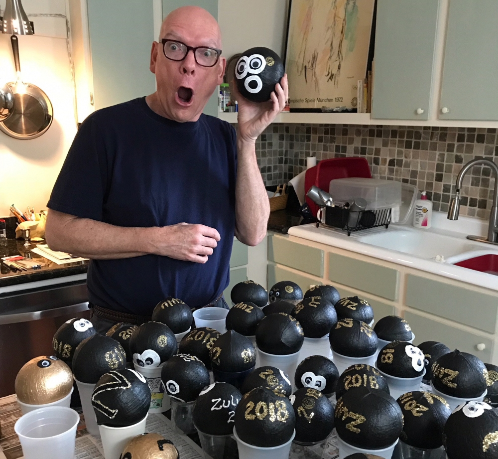 A person in a kitchen excitedly holds a black spherical object with googly eyes. The counter is covered with similar decorated objects in cups, each labeled with a year or design. The kitchen features white cabinets and a mosaic tile backsplash.