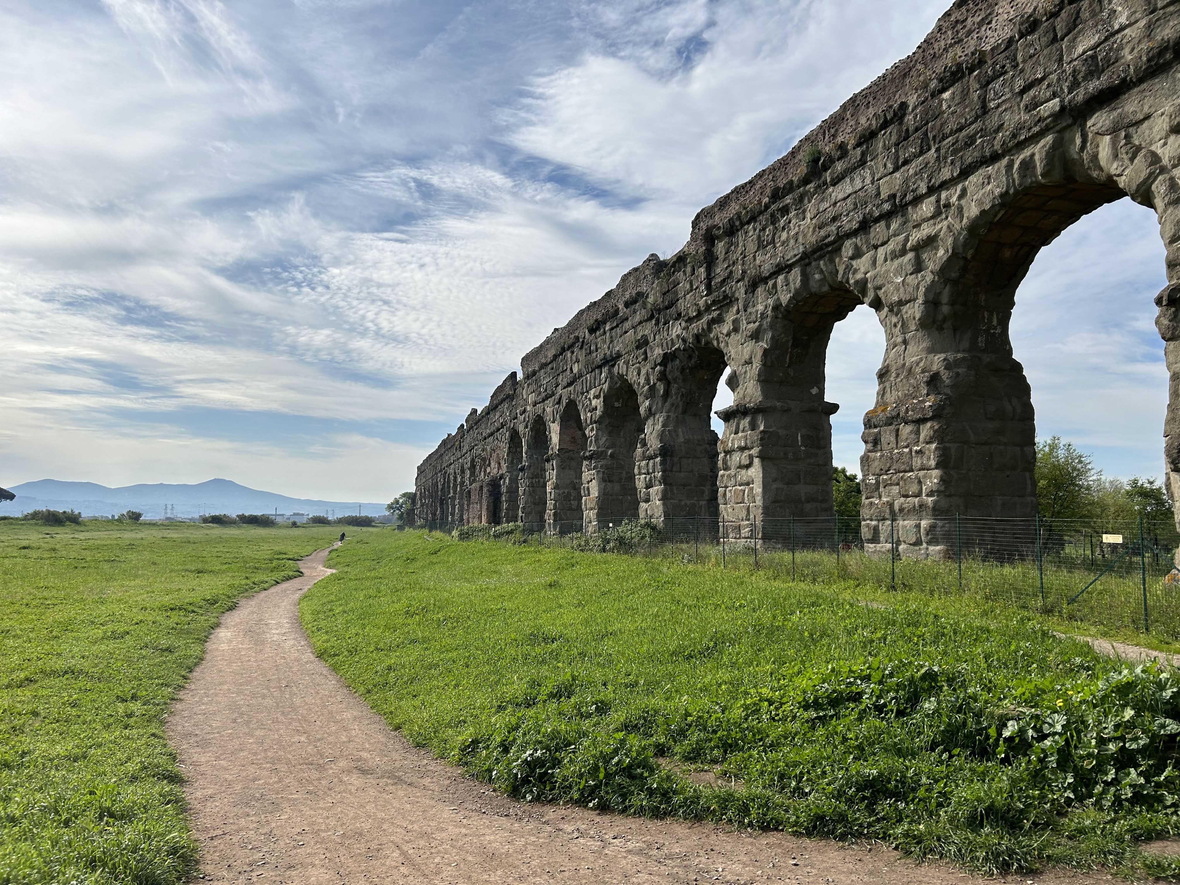 A historic stone aqueduct with arched openings stretches across a grassy landscape under a partly cloudy sky. A dirt path leads towards the structure, with mountains visible in the distant background.