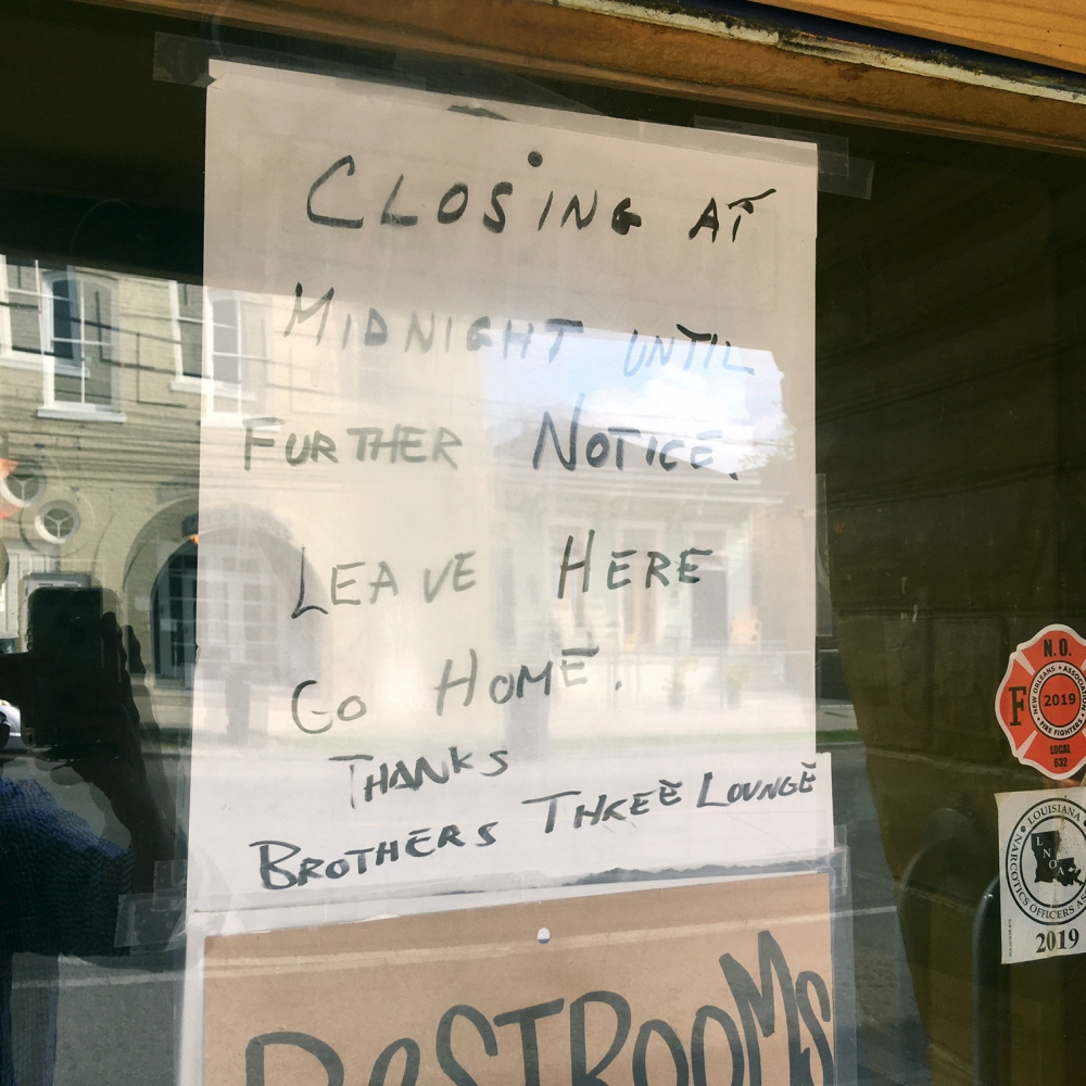 A hand-written sign taped inside a window reads: Closing at midnight until further notice. Leave here. Go home. Thanks, Brothers Three Lounge. Reflections of buildings and a person are visible in the glass.