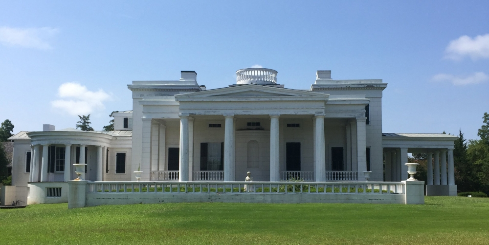 A large, white neoclassical mansion with tall columns and a central pediment. The building has multiple chimneys and a circular railing on the roof. It is situated on a grassy lawn under a clear blue sky.